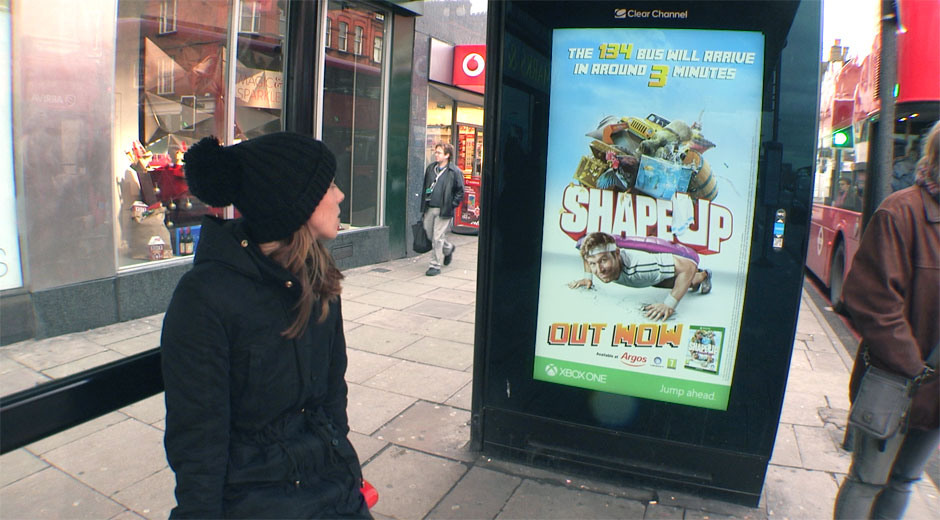 A young woman looks at a Ubisoft Shape Up dynamic digital out of home poster whilst waiting for a bus